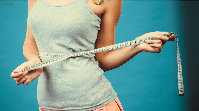 Woman wrapping a tape measure around her waist against blue backdrop.